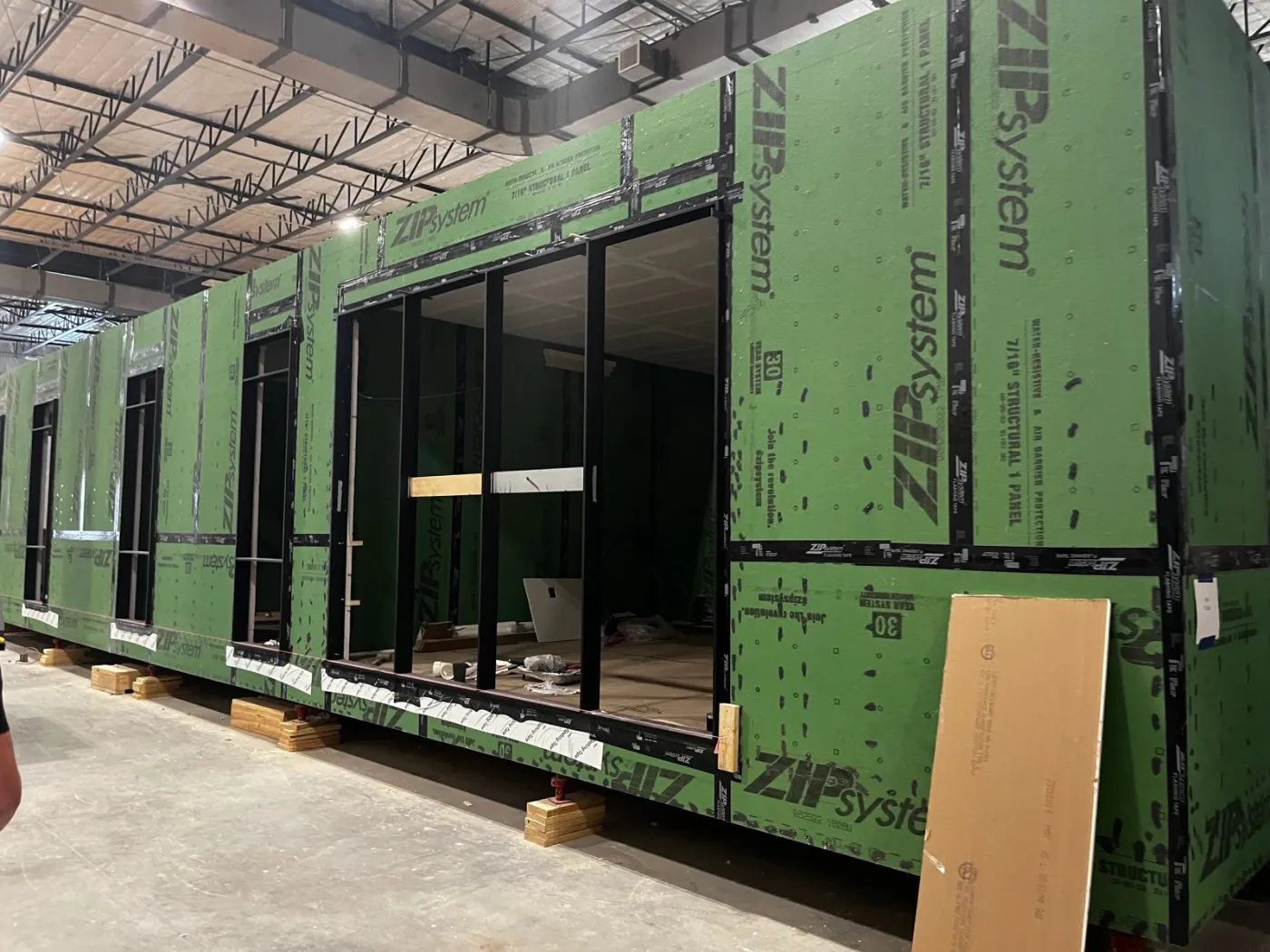 A module wrapped in green sheathing, with large window openings, on the factory floor