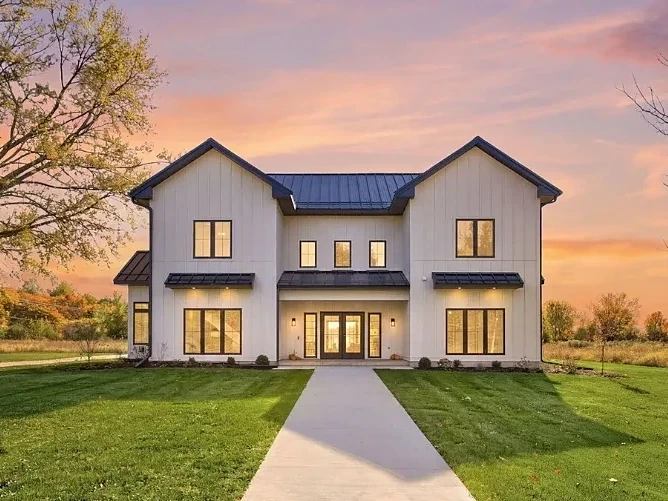 Completed two-story white board-and-batten home with a metal roof and lit windows at sunset, set in open fields