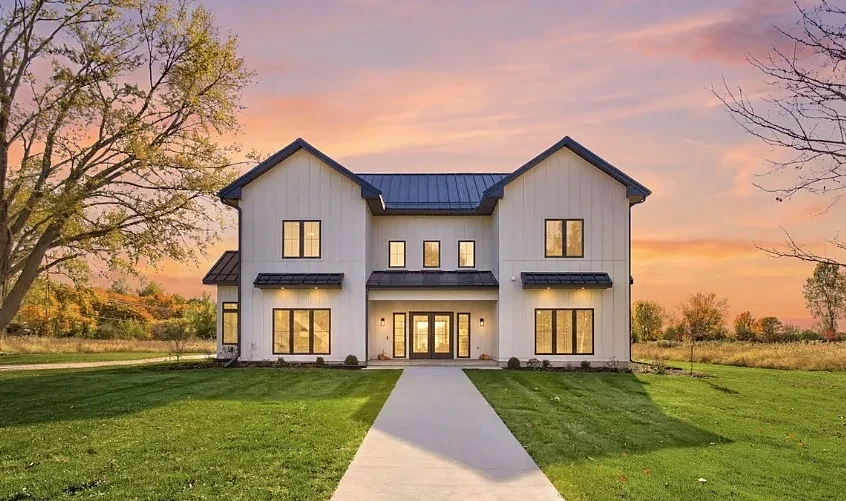Completed two-story white board-and-batten farmhouse with a metal roof and lit windows at sunset, set in open fields