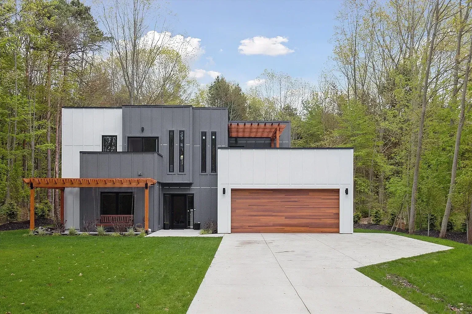 Modern two-story home with white and charcoal panel siding, a cedar garage door, and cedar pergolas on a wooded lot