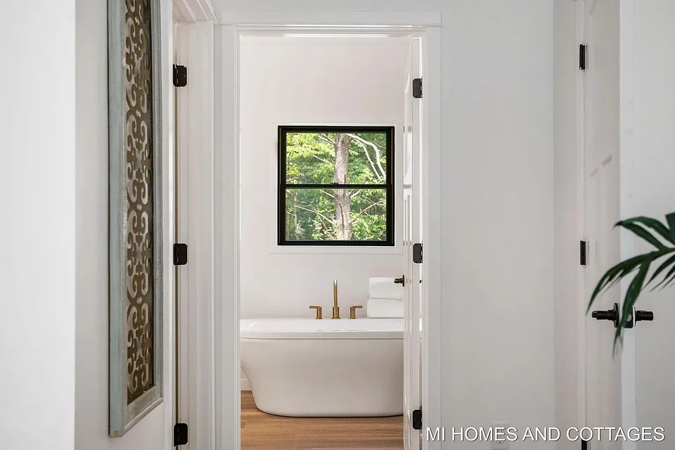 View down the hall into the primary bath, with a freestanding tub and brass fixtures under a window