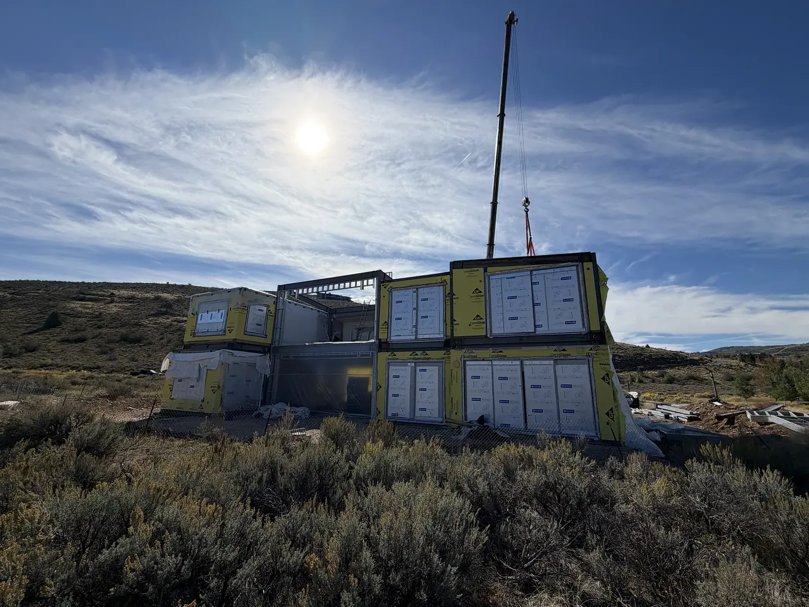 Stacked steel modules on a hillside build site under a bright sky, with a crane behind them