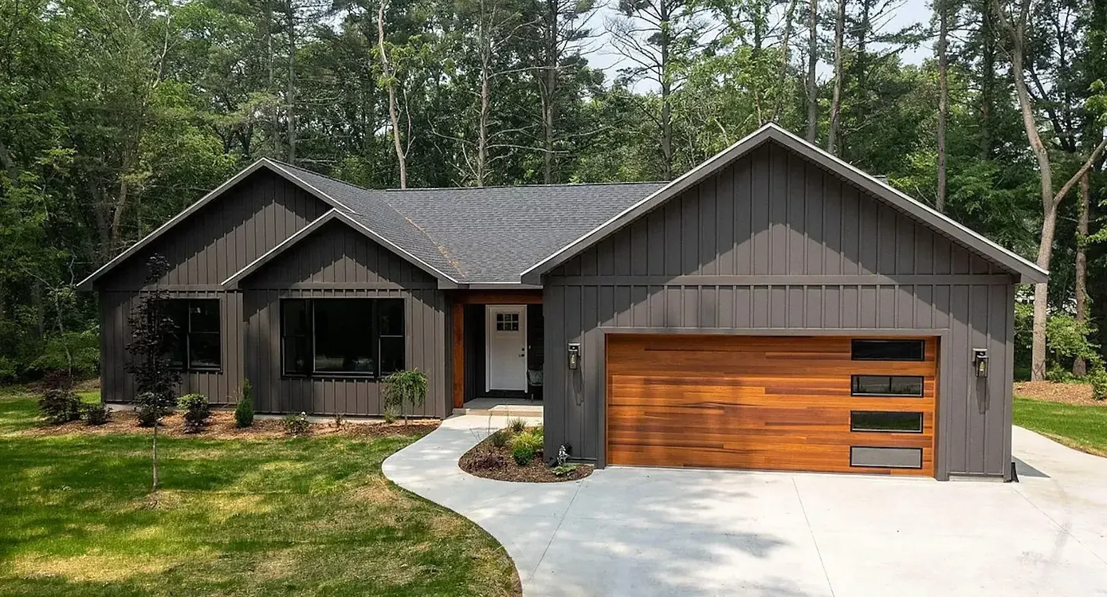 Single-story home with dark board-and-batten siding and a cedar garage door in a wooded setting