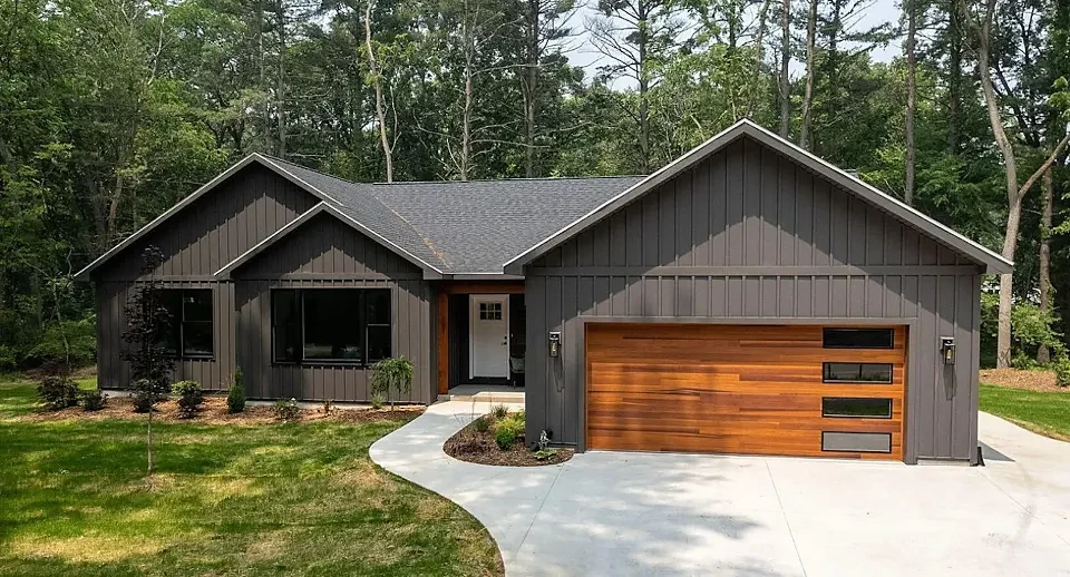 Completed single-story home with dark board-and-batten siding and a cedar garage door in a wooded setting