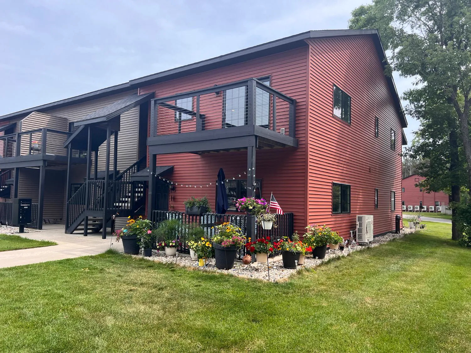 Twin Oaks building corner with red and tan lap siding, an upper balcony with cable railing, and a planted patio